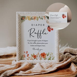 May include: A white frame with a floral design and a sign that says "DIAPER Raffle". The sign says "If you brought a pack of diapers for the raffle, please place your ticket and diapers here. Thank you". There is a small card with the text "Diaper Raffle" and a ladybug design.