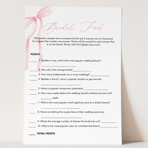 May include: A white bridal shower game card with pink accents. The card features the text "Bridal Feud" and includes fill-in-the-blank questions about weddings. A pink ribbon design is in the upper left corner.