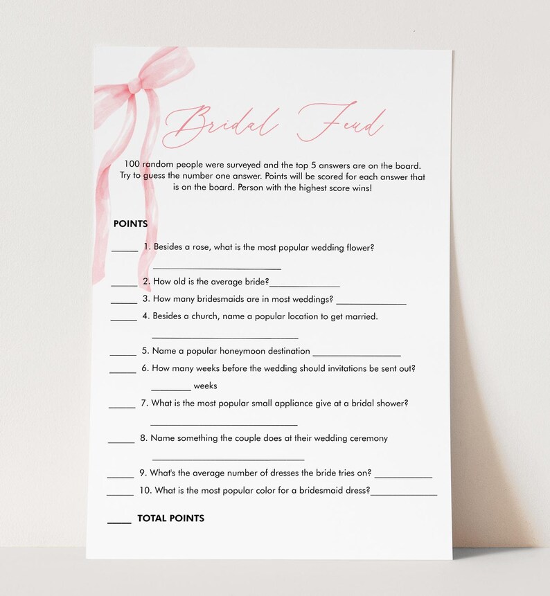 May include: A white bridal shower game card with pink accents. The card features the text "Bridal Feud" and includes fill-in-the-blank questions about weddings. A pink ribbon design is in the upper left corner.