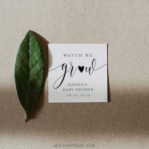 Puede incluir: Una invitaci&oacute;n cuadrada y blanca para baby shower con el texto "Watch Me Grow" en escritura elegante, un coraz&oacute;n negro y los detalles "Hanna's Baby Shower" y la fecha. Una hoja verde descansa junto a la invitaci&oacute;n sobre un fondo neutro.