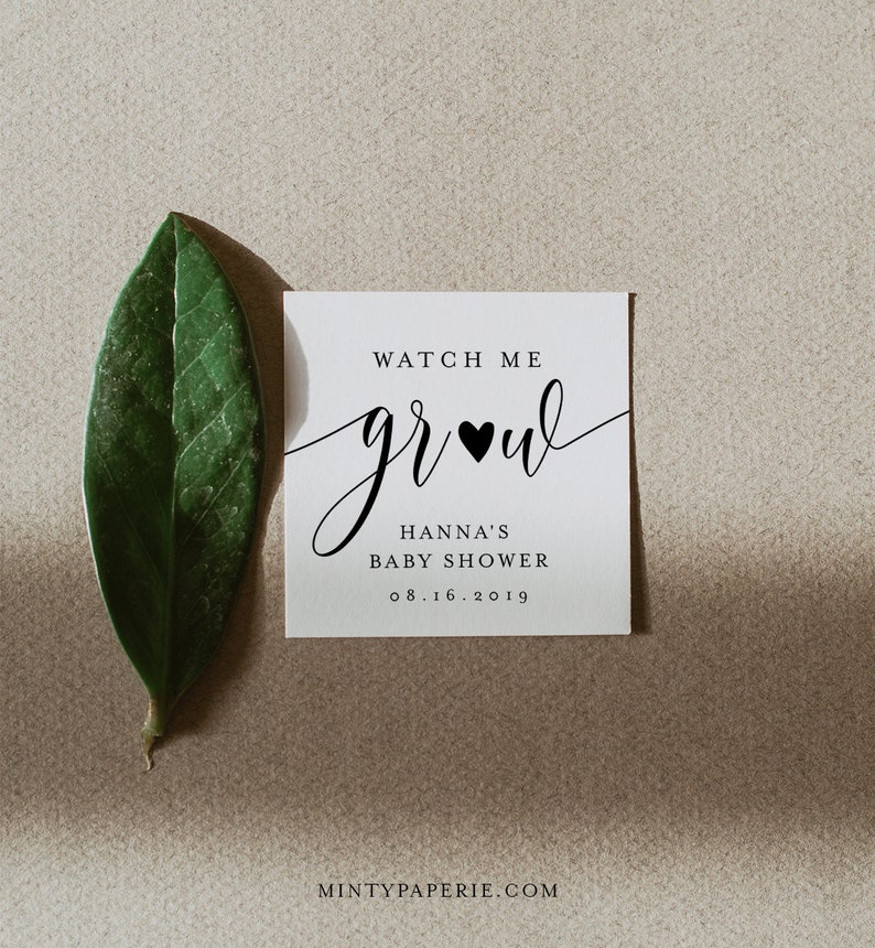 Puede incluir: Una invitaci&oacute;n cuadrada y blanca para baby shower con el texto "Watch Me Grow" en escritura elegante, un coraz&oacute;n negro y los detalles "Hanna's Baby Shower" y la fecha. Una hoja verde descansa junto a la invitaci&oacute;n sobre un fondo neutro.