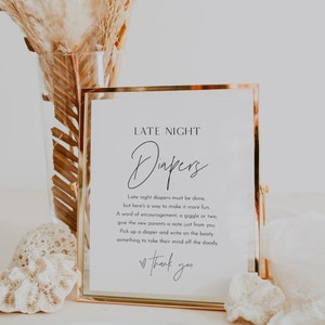 May include: A white sign with a gold frame, with the text "LATE NIGHT Diapers" and instructions for guests to write notes on diapers for the new parents. The sign is sitting on a table with a vase of dried flowers.