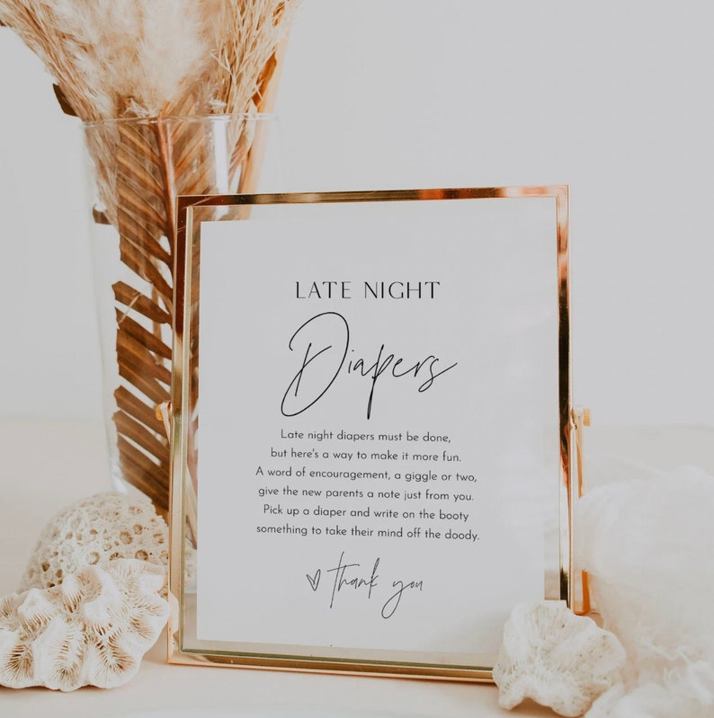May include: A white sign with a gold frame, with the text "LATE NIGHT Diapers" and instructions for guests to write notes on diapers for the new parents. The sign is sitting on a table with a vase of dried flowers.