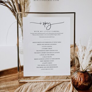 May include: A printable wedding sign with a gold frame, featuring the text "i spy" and a list of photo prompts for guests to capture at the wedding. The sign is on a wooden table with a beige fabric and a vase with dried flowers.