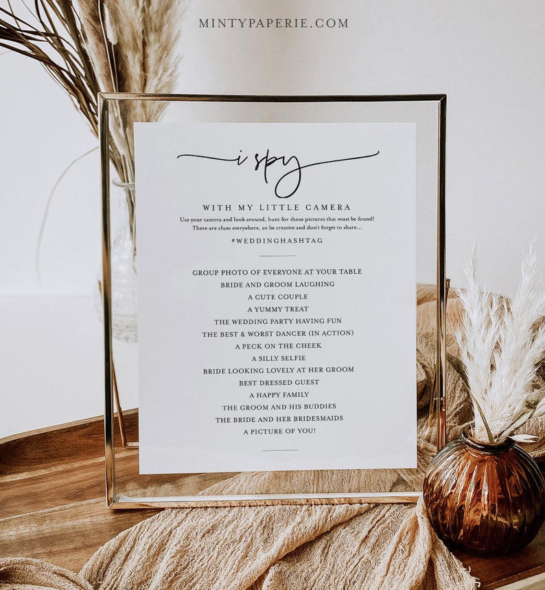 May include: A printable wedding sign with a gold frame, featuring the text "i spy" and a list of photo prompts for guests to capture at the wedding. The sign is on a wooden table with a beige fabric and a vase with dried flowers.