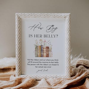 May include: A white frame with a sign that says "How Big Is Her Belly?" with a stack of books and flowers. The sign also says "Pull the ribbon and cut where you think will fit around the mommy-to-be's belly. The person with the ribbon cut closest to her actual size wins! Good luck."