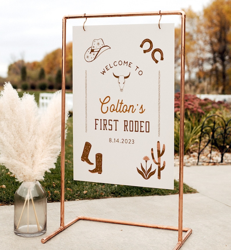 May include: A white sign with brown text and illustrations announcing "Welcome to Colton's First Rodeo" with the date 8.14.2023. The sign is on a copper stand.