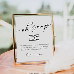 May include: A white sign with black text and a camera icon that says "oh snap" and "Use these cameras to capture special moments tonight. Take a few snaps, then pass it on and leave it on the table once the film runs out. Sophia and James". The sign is in a gold frame and sitting on a wooden table.