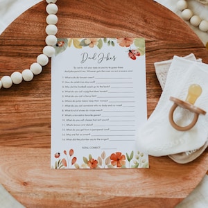 May include: A printable game card with a floral border and a title that reads "Dad Jokes". The card features a list of 14 dad jokes with blank lines for players to write in their answers.