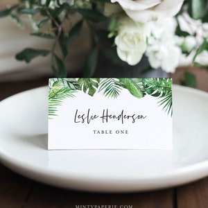 May include: White place card with a green tropical leaf border. The text reads "Leslie Henderson, Table One".