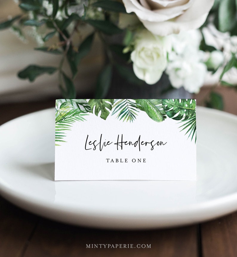 May include: White place card with a green tropical leaf border. The text reads "Leslie Henderson, Table One".