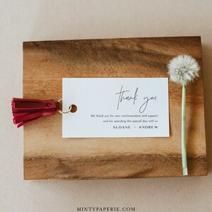May include: A white thank you card with black text that reads "thank you" in cursive. The text below reads "We thank you for your continuous love and support and for spending this special day with us. SLOANE + ANDREW". The card is on a wooden surface with a red tassel attached to the top left corner.