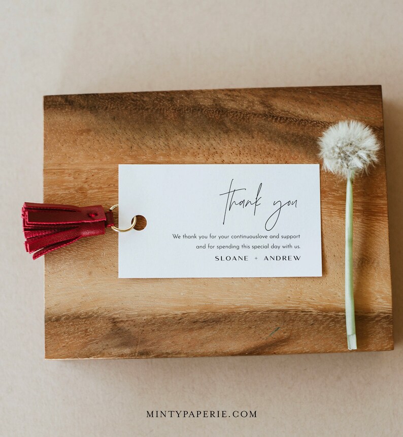 May include: A white thank you card with black text that reads "thank you" in cursive. The text below reads "We thank you for your continuous love and support and for spending this special day with us. SLOANE + ANDREW". The card is on a wooden surface with a red tassel attached to the top left corner.