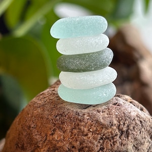May include: A tower of six pieces of sea glass in shades of blue, white, and green, carefully arranged on a piece of driftwood. The sea glass has a smooth, frosted texture. The background features blurred green foliage.