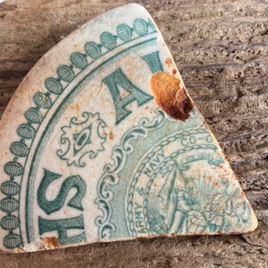 Może przedstawiać: Fragment ceramiczny z wyblakłymi turkusowymi i białymi detalami. Widoczny jest tekst "SH" oraz słowa "ARMY & NAVY CO-G". Projekt zawiera ozdobne obramowania i centralny emblemat, wykazujący oznaki zużycia.