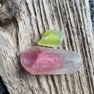 以下が含まれることがあります： ピンクとグリーンのシーグラス2個が、風化した流木の上に置かれています。ピンク色の大きなシーグラスにはハート型のくぼみがあります。緑色のシーグラスは小さく、より角張った形をしています。