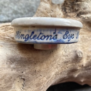May include: A small, off-white ceramic bottle stopper with blue lettering that reads "Singleton's Eye Ointment." The stopper has a rounded top and a cylindrical base. The background is a piece of driftwood.
