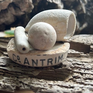 May include: A collection of weathered, off-white ceramic pieces, including a small cylinder, a sphere, and a cup-shaped object, arranged on a piece of driftwood. The base of the arrangement has the text "CO ANTRIM" in black lettering.