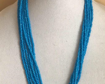 Könnte beinhalten: Eine mehrsträngige Halskette aus kleinen, runden, leuchtend blauen Perlen. Die Halskette wird auf einer weißen Mannequin-Büste präsentiert. Die Stränge sind hinten mit einem dunklen Verschluss verbunden.