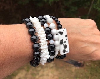 Könnte beinhalten: Eine Reihe von Armbändern mit schwarzen und weißen Perlen. Die Armbänder umfassen große, glänzende schwarze Perlen, weiße muschelartige Stücke und rechteckige Perlen mit weißen und schwarzen Flecken. Die Armbänder werden am Handgelenk getragen.