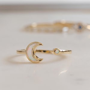 May include: A gold-colored ring featuring a crescent moon design with a white, iridescent inlay. A small, round, white stone is set on the band. The ring is displayed on a white surface, with other rings slightly out of focus in the background.