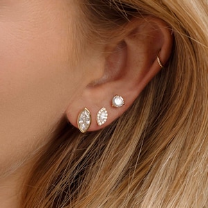 Op de afbeelding: Close-up van een oor versierd met drie gouden oorbellen, elk met een sprankelende edelsteen. Een gouden ketting met kleine edelstenen en een hanger is ook zichtbaar.