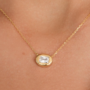 Può includere: Una collana dorata con un pendente rettangolare in pietra preziosa trasparente. Il pendente è incastonato in una lunetta dorata e pende da una delicata catena d'oro. La collana è mostrata su uno sfondo bianco.