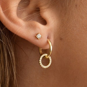 May include: Gold hoop earrings with a smaller circle charm, both adorned with small sparkling stones. The earrings are worn in a person's ear.