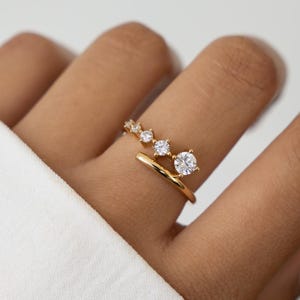 Può includere: Un anello dorato con un design aperto unico, caratterizzato da una serie di pietre preziose chiare e scintillanti. L'anello ha uno stile moderno e minimalista, con pietre preziose di dimensioni diverse. L'anello è indossato su un dito.