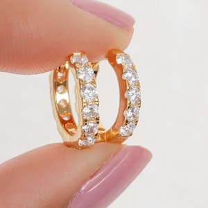 May include: A pair of gold-coloured hoop earrings, each set with a row of small, clear gemstones. The earrings are held between two fingers against a soft, neutral background. The gemstones sparkle, catching the light.