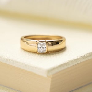 May include: A gold ring with a rectangular, clear gemstone set in the center. The ring is resting on a book with cream-colored pages. The ring's band is thick and polished, reflecting the light.