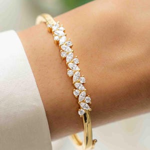 May include: A gold-colored bracelet with a floral design. The bracelet features a series of clear, sparkling stones set in a leaf-like pattern. The bracelet is worn on a wrist, and the background is blurred.