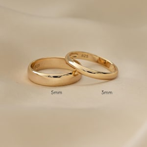 May include: Two gold rings, one 5mm wide and the other 3mm wide, are displayed on a cream-colored fabric. The rings have a polished finish and are set against a soft, neutral background. The rings are likely wedding bands or fashion accessories.