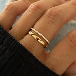 Puede incluir: Dos anillos de oro se muestran en un dedo. Uno es una banda lisa y pulida. El otro es una banda delgada adornada con pequeñas piedras brillantes. Los anillos se colocan sobre un fondo de manga negra y mezclilla azul.