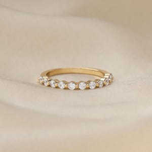 May include: A gold ring featuring a line of small, clear gemstones. The ring is displayed on a soft, neutral backdrop, accentuating the sparkle of the stones. The design is simple and elegant, suitable for everyday wear or special occasions.