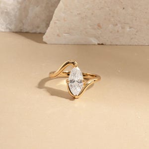 May include: A gold ring with a marquise-cut diamond. The ring has a unique, flowing design with a delicate band that curves around the finger. The diamond is set in a prong setting, and the ring is displayed on a neutral background.