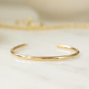 May include: A gold-colored open cuff bracelet. The bracelet is a simple, minimalist design with a smooth, polished finish. The jewelry is displayed on a white marble surface.