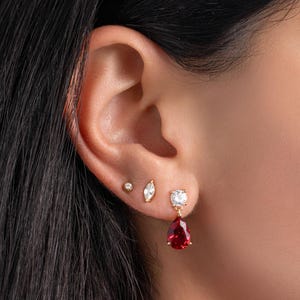 May include: A set of three gold-tone earrings worn in a single ear. The set includes a small round stud, a marquise-cut stud, and a drop earring featuring a round clear stone at the top and a vibrant red pear-shaped stone dangling below.