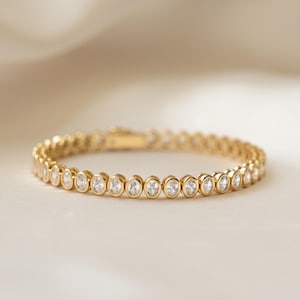 May include: A gold bracelet with a series of round, clear gemstones set in individual bezels. The bracelet is displayed on a soft, off-white surface, with a blurred, light-colored background. The bracelet is a jewelry item.