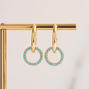 May include: A pair of gold-colored hoop earrings. Each earring features a circular design with small, light blue gemstones encircling the inner ring. The earrings are displayed on a gold-colored jewelry stand against a white background.