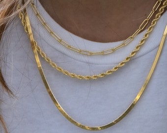 May include: Three gold necklaces layered on a person's neck. The necklaces are a thin chain, a thicker rope chain, and a flat chain.