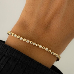 Op de afbeelding: Een gouden armband met kleine ronde, heldere stenen die in het metaal zijn gezet. De armband zit om een pols.