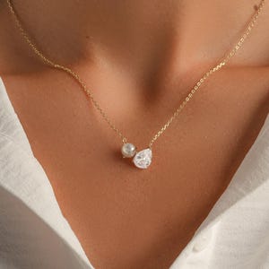May include: A delicate gold necklace featuring a small pearl and a pear-shaped, clear crystal pendant. The necklace is worn against a white collared shirt.
