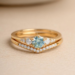 May include: A gold engagement ring set featuring a light blue gemstone center stone and a matching band. The main ring has a round, light blue center stone, surrounded by smaller white gemstones. The band is a V-shaped design, also adorned with small white gemstones.