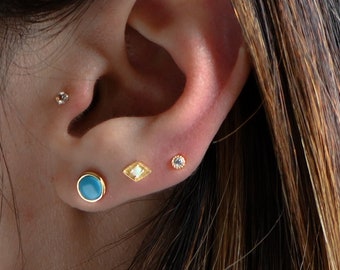 May include: A close-up of a person's ear with three small earrings. The earrings are a turquoise stone, a gold diamond shape with a white stone in the centre, and a small white stone.