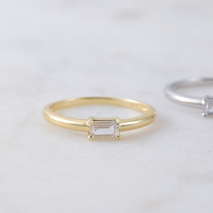 May include: Two delicate rings, one gold and one silver, each featuring a rectangular, clear gemstone. The rings are set against a white marble surface, creating a minimalist and elegant aesthetic. The gold ring has a warm, yellow hue, while the silver ring has a cool, metallic sheen.