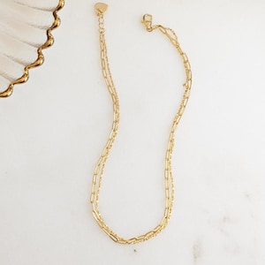 Puede incluir: Un collar de cadena de oro con un diseño delicado y en capas. El collar presenta dos cadenas de diferentes grosores, creando un look elegante y moderno.