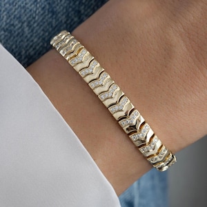 May include: A gold-toned bracelet with a chevron design, set with small, sparkling stones. The bracelet is worn on a wrist, against a backdrop of a white shirt and denim fabric.