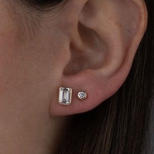 Puede incluir: Un par de pendientes de oro con piedras preciosas transparentes. Un pendiente tiene una piedra preciosa de forma rectangular, mientras que el otro pendiente tiene una piedra preciosa redonda.