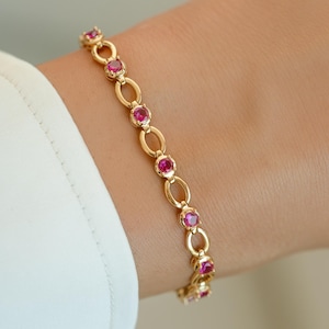 May include: A gold chain bracelet featuring a series of alternating oval links and round, bezel-set vibrant pink gemstones. The bracelet is worn on a wrist, showcasing the polished metal finish and the consistent spacing of the circular pink stones.
